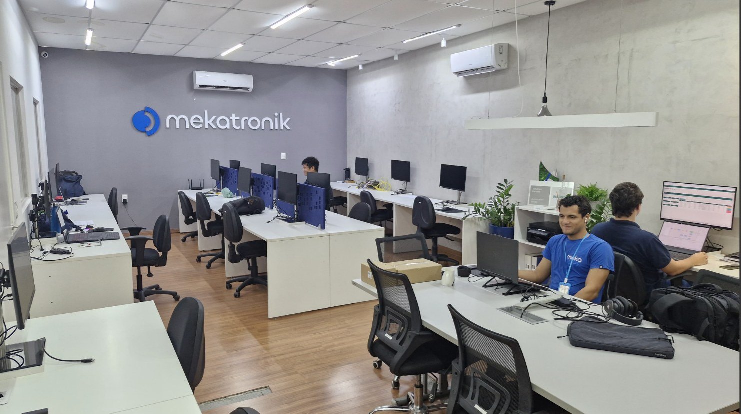 Escritório de engenharia da Mekatronik: fileiras de estações de trabalho com monitores, um engenheiro analisando uma planilha de produção em tela e a marca Mekatronik aplicada na parede ao fundo.